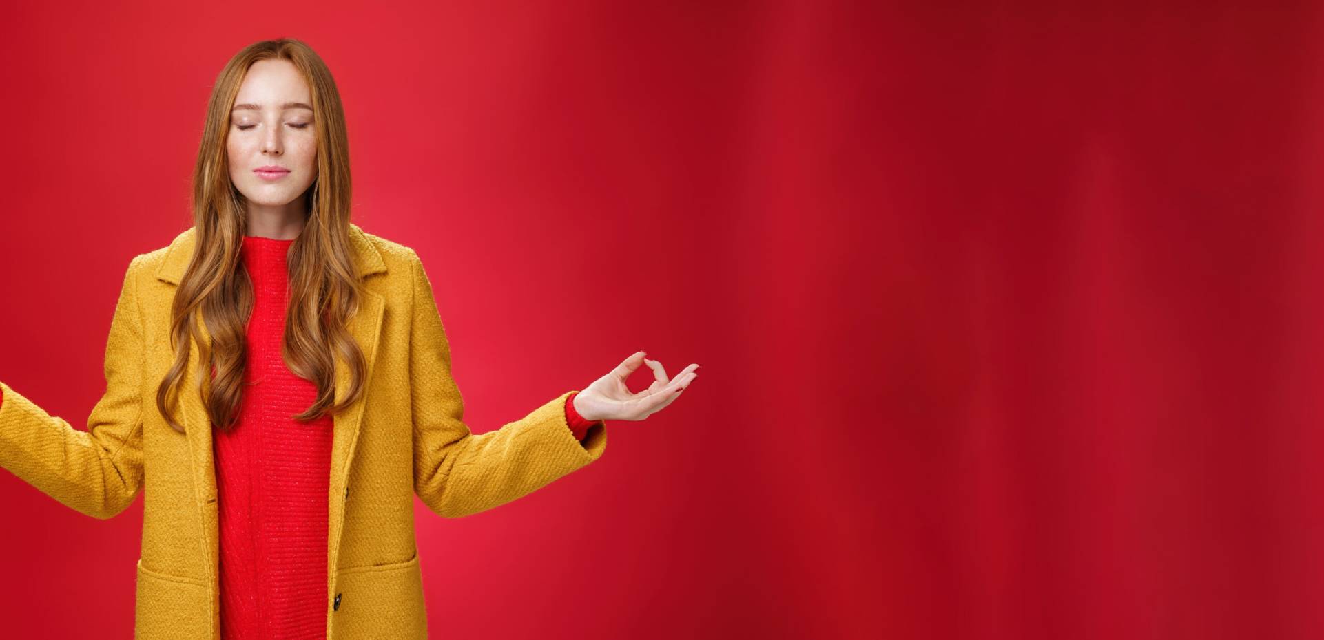 girl keep calm releasing stress with meditation, posing in yellow coat, close eyes and looking relieved as extending hands sideways with mudra gesture, doing yoga against red background in lotus pose